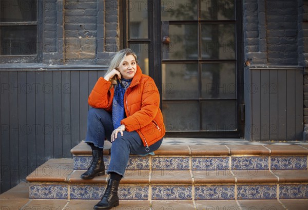 Happy smiling middle aged woman in orange down jacket sitting on concrete stairs outdoors and