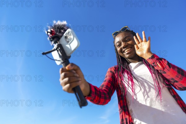 African woman waving while vlogging using mobile phone and microphone outdoors