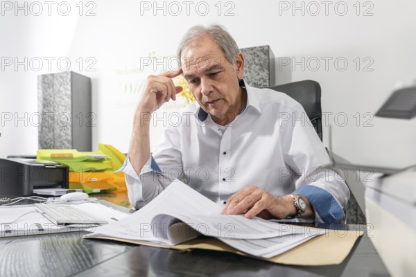 Elderly man in a white shirt sits at a desk and leafs through a document file