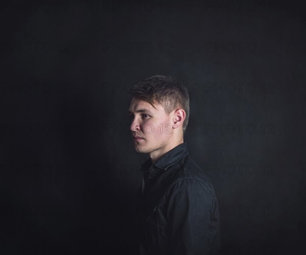 Thoughtful young man side view portrait, looks upset and tired against a dark wall background.