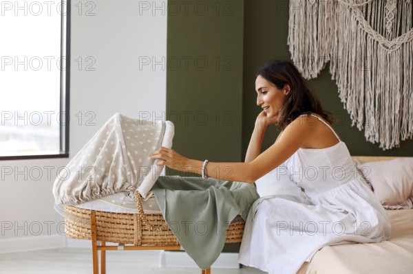 Young beautiful happy woman mother looking at her little newborn baby sleeping in bed bassinet at