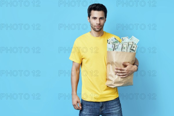Handsome man hold paper bag full of stacks of money isolated on blue background. Success, business,