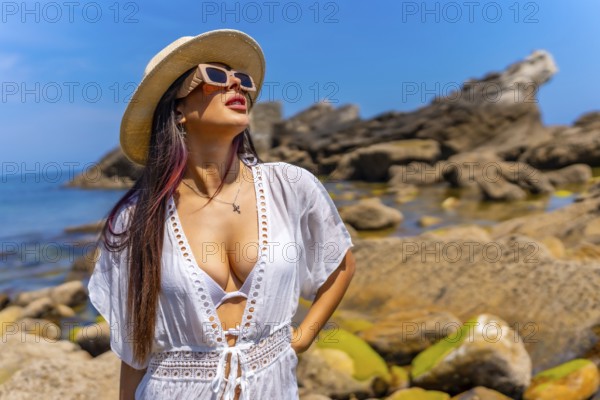 A woman wearing a white dress and a straw hat is standing on a beach. She is looking out at the