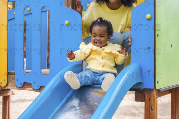 Baby girl sliding in a blue slide in a park with her mother in her back watching and controlling
