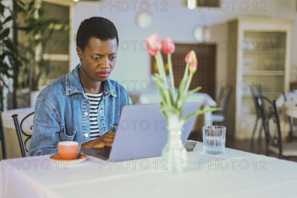 Woman using laptop with coffee cup and tulips on a table in sunlit restaurant