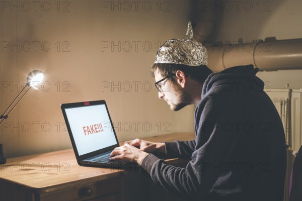 Young man with aluminum cap is sitting in the dark basement in front of a laptop. Conspiracy theory