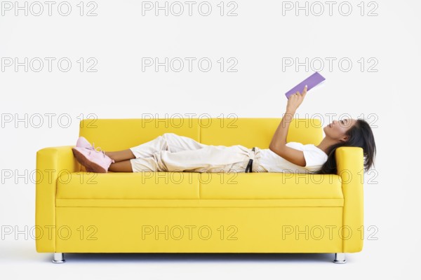 Pretty asian woman reading book lying at cozy yellow couch, concepts of home and comfort, hobby,