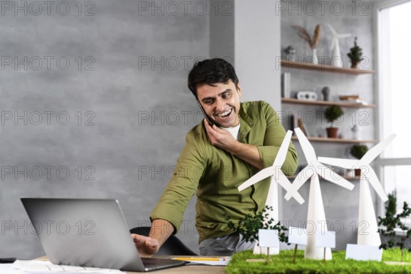 Smiley man working eco friendly wind power project while talking phone using laptop