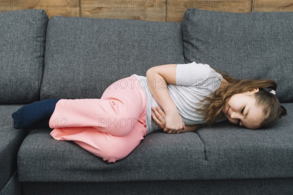 Little girl suffering from stomach ache lying gray sofa