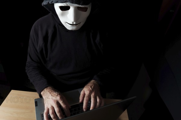 Hacker man dressed in black, hooded with mask typing on a computer cybercrime concept