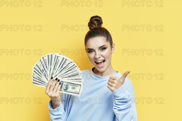 Happy excited young woman showing money banknotes with thumbs up on yellow background. Motivation,
