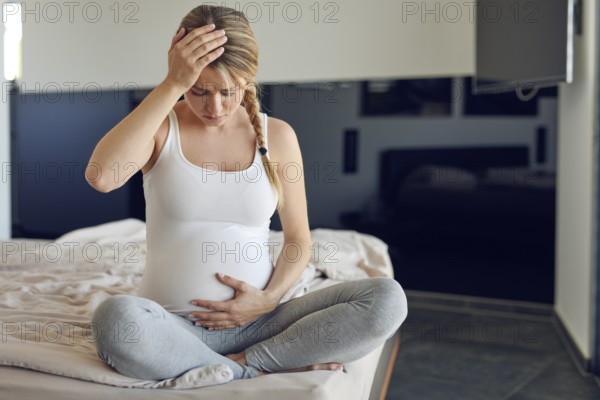 Troubled heavily pregnant young woman sitting cross legged on a bed cradling her swollen abdomen