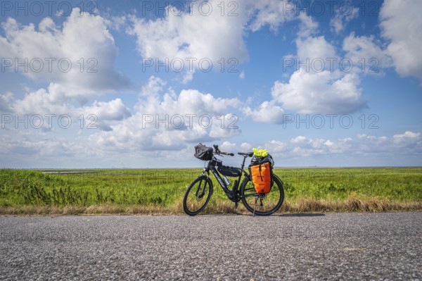 A loaded touring bike stands at the roadside with a wide field and open sky in the background,