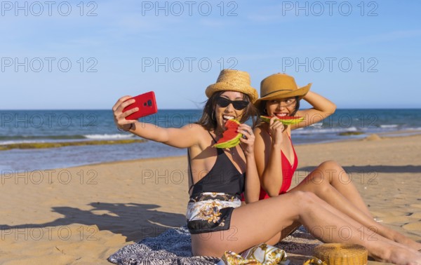 Friends on the beach in summer eating a watermelon with the sea in the background, taking a selfie