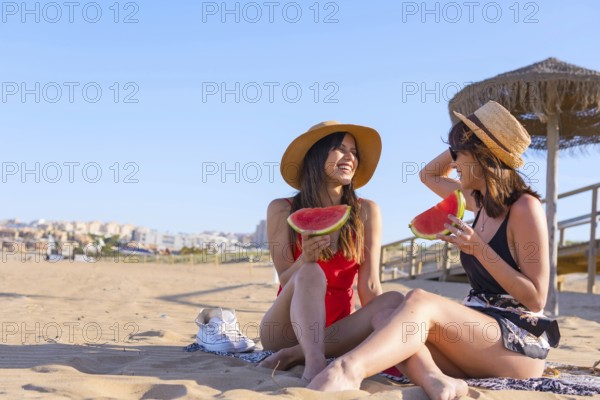 Female friends on vacation at the beach in summer eating a watermelon, sitting on the sand next to