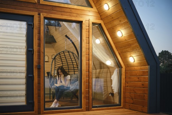 Woman relaxing in a hanging chair with a book inside a stylish wooden cabin. The warm lighting and