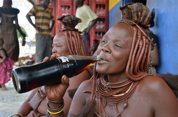 Traditionally dressed Himba woman drinking a coke in front of a bar, Opuwo, Kaokoland, Kunene,
