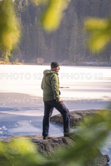 Person standing on the shore of a frozen lake, framed by rocky shore and forest, Glaswaldsee, Bad