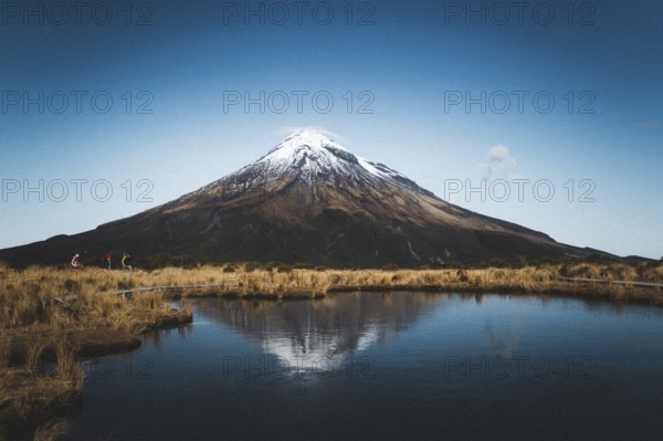 A snow-capped mountain is reflected in a still lake while people walk along the shore, Mount