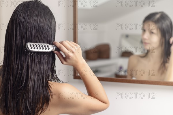 Young woman standing looking at her reflection in the bathroom mirror while brushing her wet hair