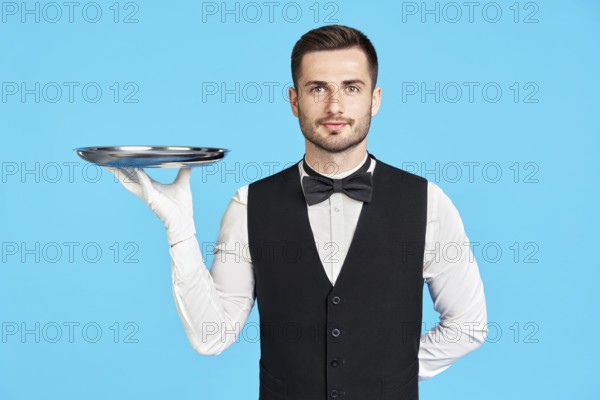 Attractive young waiter holding empty silver tray over blue background with copy space. Restaurant,