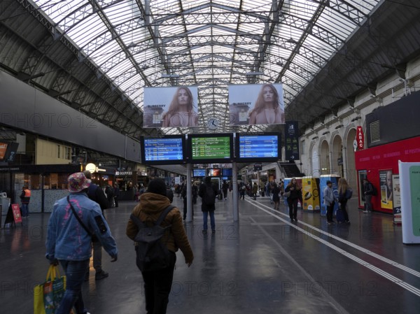 Travellers in a station concourse under a large glass-covered ceiling with timetable displays and