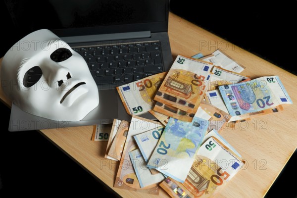 On a laptop there is a white mask and a pile of euro banknotes, cybercrime concept