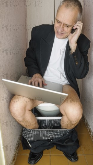 A workaholic adult businessman is using the computer laptop and the mobile phone, also called