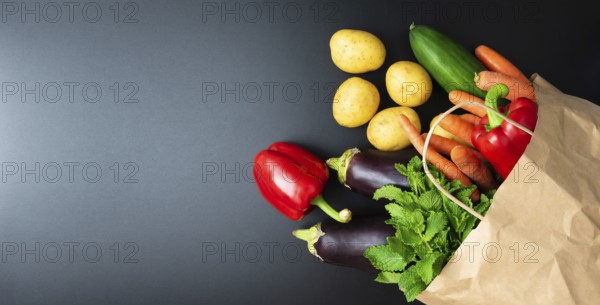 above view of fresh organic vegetables spilling out of paper shopping bag on dark kitchen counter,