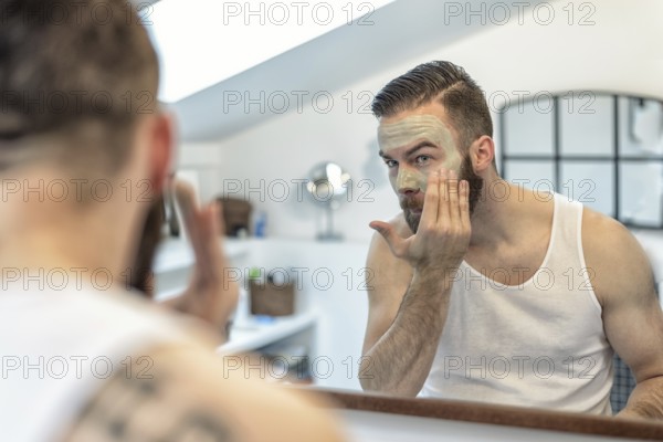 Reflection in the bathroom mirror of a young bearded man applying a face mask to his skin in a