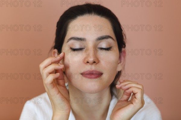Positive young brunette female with eye patches touching her face and smiling during skin care