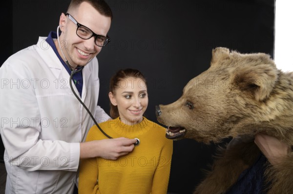 Smiling young doctor with stethoscope and patient examining bear, studio medical humor of whom to