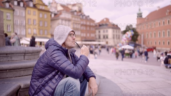 Portrait of adult man with glasses sitting in the hood on square and smoking a tobacco pipe in the