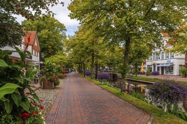A paved path next to a canal, flanked by lush plants and residential buildings, Papenburg, Emsland,