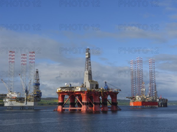 Three oil rigs standing side by side in a calm sea under a blue sky, partly cloudy, drilling rigs