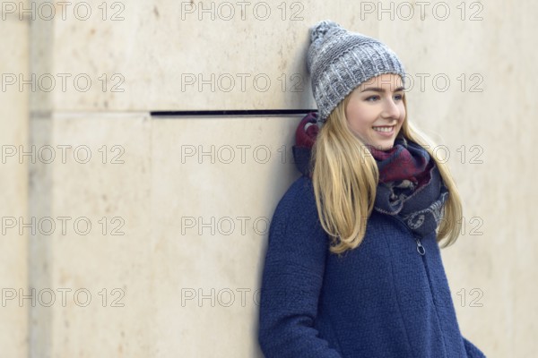 Smiling attractive young blond woman in a warm blue winter outfit standing daydreaming leaning back