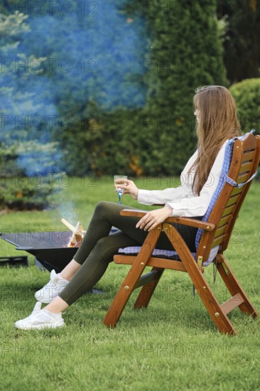 Young woman resting in lounge chair on backyard next to a barbecue, view on profile