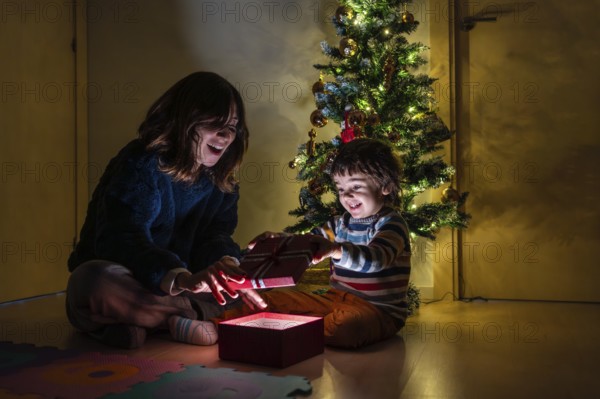A woman and a child are opening a present in front of a Christmas tree. Scene is joyful and