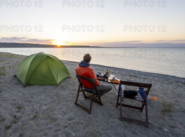 Camping at the lakeshore, young man sitting next to tent on camping chair during dinner, sunset at