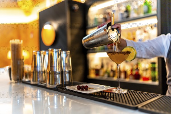 Hand of a waiter filling cocktail into martini glass garnished with lemon in the counter of a