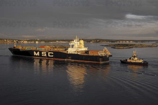 A cargo ship and a tugboat sail on calm water at sunset, loaded with containers, Gothenburg, Baltic