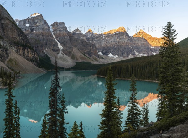 Sunset over a serene lake surrounded by rocky mountains and pine trees, with reflections in the