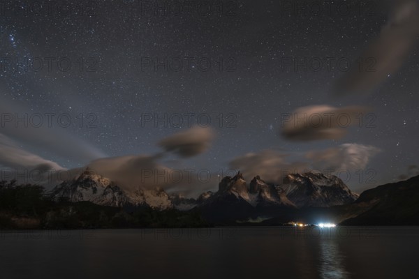 Night sky filled with stars over snow-capped mountains and a calm lake, with clouds and reflections
