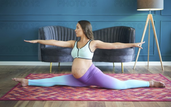 Pregnant sporty woman practicing yoga at home doing stretching exercise the side split pose