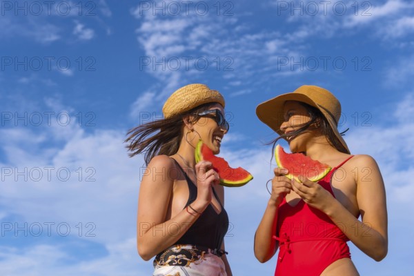 Female friends at the beach in summer eating a watermelon with the sky in the background, shot from