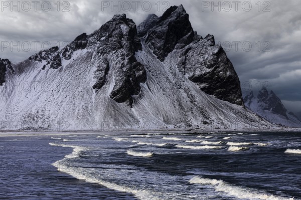 White foamy waves on the beach of Stokkness, behind snowy rocky slopes of Vestrahorn, near Höfn,
