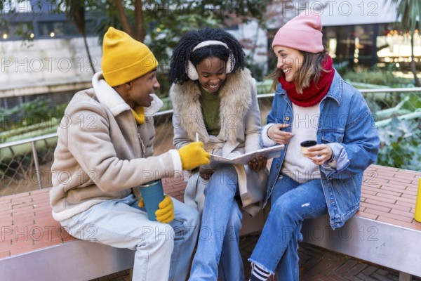 Three cheerful young students are sitting on a bench, studying and having a coffee break outdoors