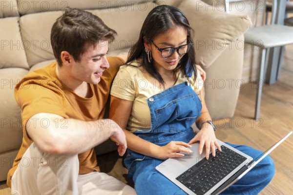 Young multiethnic couple sitting on floor and using laptop, browsing internet and smiling in cozy