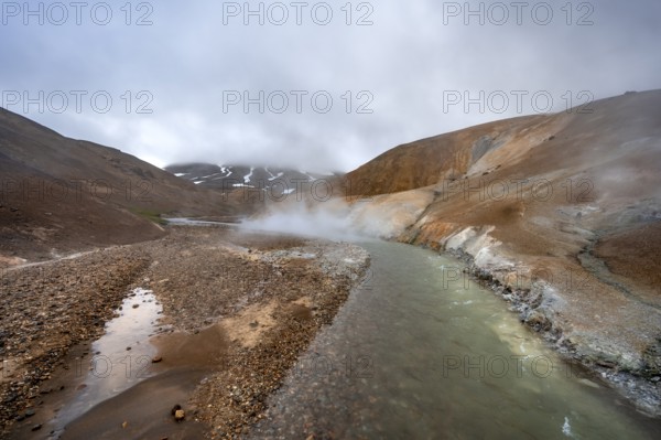 Steaming stream between colourful rhyolite mountains with snowfields, Hveradalir geothermal area,