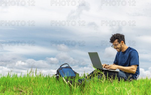 Smiling man with laptop sitting on the grass in the field. Side view of man sitting on green grass